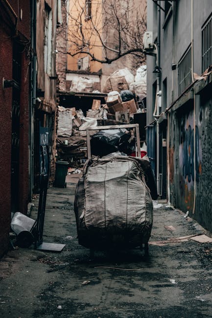 A narrow urban alleyway filled with various types of waste and rubbish. In the foreground, a large black trolleypulled by a long metal handle is covered with a grey, weathered fabric material, placed on a concrete ground that appears stained and uneven. To the left side of the alley, a small white container lies on the ground, alongside scattered debris. The right side features a grey building with metal security bars on the windows and faded graffiti artwork. In the background, a heap of discarded cardboard boxes, plastic bags, and other waste materials is piled against a wooden fence and the rear facing walls of buildings, with some items leaning or stacked haphazardly. Bare tree branches are visible above the waste heap, and the overall scene is dimly lit, suggesting an overcast day or late afternoon atmosphere. This setting signals an environment where independent rubbish removal or private waste collection services might operate outside standard municipal waste disposal routes, as exemplified by Rubbish Removal Islington.