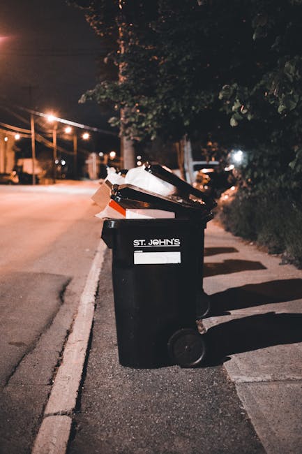 A narrow urban alleyway filled with various types of waste and rubbish. In the foreground, a large black trolleypulled by a long metal handle is covered with a grey, weathered fabric material, placed on a concrete ground that appears stained and uneven. To the left side of the alley, a small white container lies on the ground, alongside scattered debris. The right side features a grey building with metal security bars on the windows and faded graffiti artwork. In the background, a heap of discarded cardboard boxes, plastic bags, and other waste materials is piled against a wooden fence and the rear facing walls of buildings, with some items leaning or stacked haphazardly. Bare tree branches are visible above the waste heap, and the overall scene is dimly lit, suggesting an overcast day or late afternoon atmosphere. This setting signals an environment where independent rubbish removal or private waste collection services might operate outside standard municipal waste disposal routes, as exemplified by Rubbish Removal Islington.