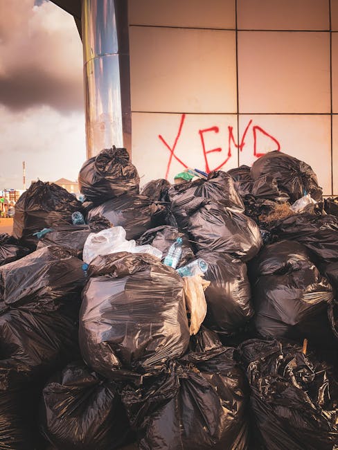 A large pile of black rubbish bags stacked against a modern building with reflective, cream-colored tiled walls. The bags are filled with waste material, with some showing signs of tearing, revealing glimpses of trash inside. Several transparent plastic water bottles with blue caps are visible among the bags, along with a few pieces of paper or plastic waste protruding from the bags. In the background, part of the urban environment can be seen under a cloudy sky, with a graffiti tag reading 'XEND' spray-painted in red on the wall behind the rubbish pile. The scene suggests an instance of unmanaged waste accumulation outside a commercial or residential property, illustrating the importance of scheduled rubbish removal services. The environment appears to be an urban sidewalk or alleyway where independent waste disposal or private collection methods, such as those offered by Rubbish Removal Islington, may be necessary to maintain cleanliness and prevent illegal dumping.