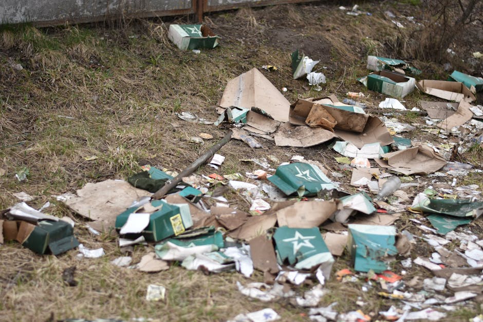 An outdoor scene shows a patch of ground next to a metallic fence, with scattered debris and litter that includes crushed and torn cardboard boxes, some of which are green with white star logos, and other paper waste. The boxes appear to be from beverage packaging, potentially beer or soda cartons, with some flattened and others partially crushed. Additionally, there are small pieces of torn paper, plastic wrappers, and other miscellaneous rubbish mixed in with the cardboard, all strewn across the grassy and dirt-covered ground. The grass is sparse, with patches of soil visible, and there are a few small dried plants or weeds near the debris. A metal pole, possibly part of a fence or utility structure, extends diagonally across the scene, partially covering some of the rubbish. The environment looks like a neglected outdoor area, potentially near an alleyway or side of a building, illustrating a typical example of rubbish that could require private waste removal services such as those provided by Rubbish Removal Islington, to clear away accumulated waste and maintain cleanliness in urban or suburban settings.