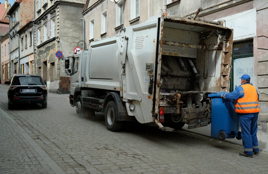 A large pile of black rubbish bags stacked against a modern building with reflective, cream-colored tiled walls. The bags are filled with waste material, with some showing signs of tearing, revealing glimpses of trash inside. Several transparent plastic water bottles with blue caps are visible among the bags, along with a few pieces of paper or plastic waste protruding from the bags. In the background, part of the urban environment can be seen under a cloudy sky, with a graffiti tag reading 'XEND' spray-painted in red on the wall behind the rubbish pile. The scene suggests an instance of unmanaged waste accumulation outside a commercial or residential property, illustrating the importance of scheduled rubbish removal services. The environment appears to be an urban sidewalk or alleyway where independent waste disposal or private collection methods, such as those offered by Rubbish Removal Islington, may be necessary to maintain cleanliness and prevent illegal dumping.