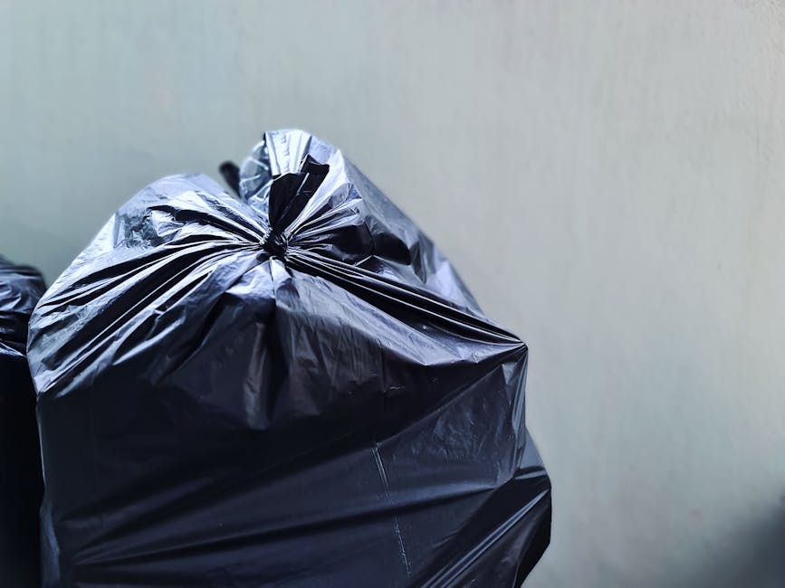 Close-up of several black plastic rubbish bags tightly knotted at the top, placed against a plain, light grey wall. The bags have a glossy, slightly crinkled texture, and are filled with waste material, causing bulges and uneven surfaces. The foreground bag is prominently positioned, occupying most of the frame, with additional bags partially visible behind it. The setting appears to be an indoor or enclosed area, possibly a storage space or a designated rubbish collection point, with no other objects or environmental features visible. The neutral lighting highlights the shiny surface of the plastic, emphasizing the bags' compacted volume. The scene reflects a typical situation where waste has been prepared for private rubbish collection or disposal, aligning with services like those offered by Rubbish Removal Islington focusing on alternative waste handling options with convenience and efficiency.