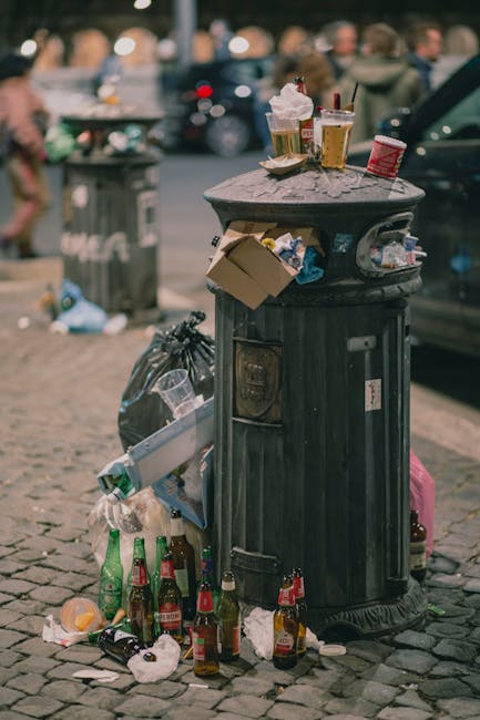 A black, vintage-style metal public trash bin with a domed, textured surface, situated on a cobblestone street. The trash bin is overflowing with mixed rubbish, including crumpled paper, discarded cups, and food packaging, some of which are visibly lidded or open. Several glass bottles, primarily green with some brown and clear, are scattered and leaning against the base of the bin, along with crumpled paper and plastic wrappers. On top of the bin are various waste items, such as used paper cups with plastic lids, a crumpled red and white disposable coffee cup, and a half-eaten sandwich in a paper wrapper, with a small plastic cup containing a beverage. In the background, blurred figures of pedestrians and parked cars are visible, along with another large bin further away. The environment appears to be an urban street scene, with natural daylight illuminating the area, demonstrating typical urban rubbish accumulation, indicative of on-site disposal or less frequent collection in a busy city setting. Rubbish Removal Islington often handles such waste on private or alternative collection schedules in the area.