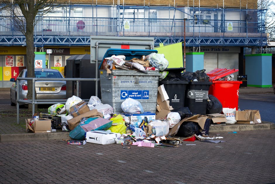 A large pile of mixed household waste and rubbish bags is overflowing from several large recycling and waste bins situated on a paved parking area. The pile includes cardboard boxes, plastic bags, paper, and discarded packaging, with some items spilling onto the surrounding pavement. The waste containers are of different colors, including a grey bin labelled for mixed paper and cardboard, a black bin, a red bin, and a green bin, all positioned closely together. Behind the rubbish, there is a building with a storefront, partially visible with signage and window displays, and a parked silver car with a UK registration plate nearby. A metal railing separates the waste area from the sidewalk, and a tree with bare branches is visible on the left side of the scene. The environment appears to be a commercial or retail district in an urban setting where improper rubbish disposal is evident, reflecting the type of scene that may require professional rubbish removal services to manage on-site clearance or private waste handling.