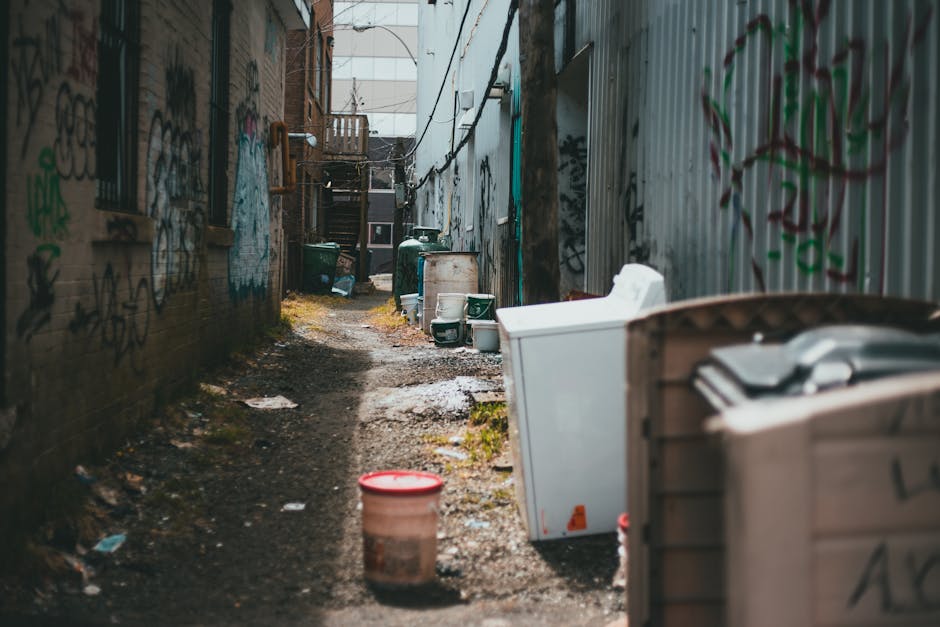 A black, vintage-style metal public trash bin with a domed, textured surface, situated on a cobblestone street. The trash bin is overflowing with mixed rubbish, including crumpled paper, discarded cups, and food packaging, some of which are visibly lidded or open. Several glass bottles, primarily green with some brown and clear, are scattered and leaning against the base of the bin, along with crumpled paper and plastic wrappers. On top of the bin are various waste items, such as used paper cups with plastic lids, a crumpled red and white disposable coffee cup, and a half-eaten sandwich in a paper wrapper, with a small plastic cup containing a beverage. In the background, blurred figures of pedestrians and parked cars are visible, along with another large bin further away. The environment appears to be an urban street scene, with natural daylight illuminating the area, demonstrating typical urban rubbish accumulation, indicative of on-site disposal or less frequent collection in a busy city setting. Rubbish Removal Islington often handles such waste on private or alternative collection schedules in the area.