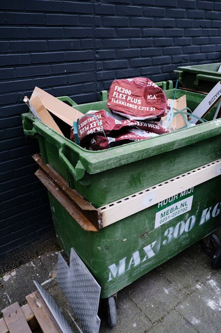 A large green wheeled waste container filled with red and white bags of building or garden supplies, including flexible hoses or tubing, situated outdoors on a paved surface beside a dark brick wall. The container has a white label with green text indicating waste management details and is positioned on a wooden pallet, with another similar container partially visible behind it. The environment appears to be a storage or back area where non-organic rubbish, possibly related to construction or renovation, is temporarily accumulated before disposal, illustrating an example of alternative waste handling outside of municipal bulky waste collection. The lighting is natural, and the scene emphasizes the practical aspects of private rubbish removal services, such as those offered by Rubbish Removal Islington, in managing bulky or non-standard waste materials.