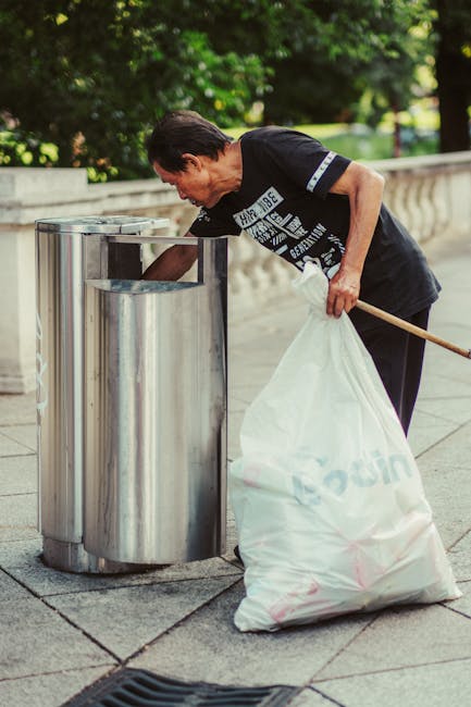 A middle-aged man with short dark hair, wearing a black T-shirt with white text and dark trousers, is seen leaning over a metal outdoor rubbish bin on a paved pavement. He is holding a large white garbage sack filled with waste, which rests on the ground beside him. In his right hand, he is gripping a wooden stick used for managing waste collection, and he appears to be in the process of depositing or removing waste from the bin. The background features lush green trees and a stone or concrete balustrade, indicating an outdoor public or park setting. The scene is illuminated by natural daylight, casting soft shadows on the ground, and the environment appears calm and orderly, reflecting typical waste disposal activities. Rubbish Removal Islington often deals with onsite waste collection and disposal, as visually represented in this urban waste management activity, demonstrating proper handling of rubbish in a public space.