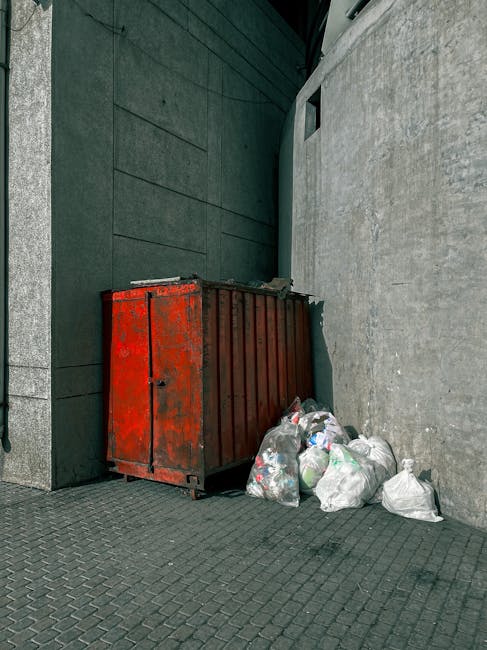 A red metal waste container with visible rust and weathering is positioned against a dark green wall on the left and a light grey concrete wall on the right, located outdoors on a paved surface. Several white plastic bags filled with rubbish are piled next to the container, some leaning against it. The bags appear to contain mixed waste materials, with some partially open, revealing contents like paper and plastic. The surrounding environment includes urban architectural elements, such as a corner where two walls meet and a small window in the dark green wall above the container. The scene is lit with natural daylight, casting soft shadows on the ground, emphasizing the textures of the metal, plastic bags, and concrete. This outdoor waste scene illustrates the type of collection needs that private rubbish removal services like Rubbish Removal Islington handle, providing an alternative to municipal waste disposal options by managing on-site clearance of mixed waste materials.