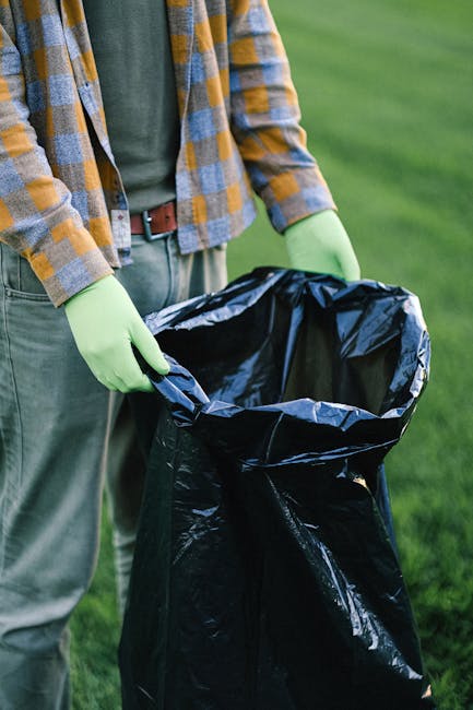 A person wearing a multicolored checkered shirt, grey trousers, and bright green gloves is holding open a large black rubbish bag, preparing to dispose of waste outdoors on a grassy area. The focus is on the individual's hands and the open rubbish bag, which appears to be made of thick plastic with a shiny, crinkled surface. The background shows a well-maintained lawn with blurred green grass, indicating an outdoor environment suitable for waste collection or clearance activities. The scene suggests an organized approach to rubbish removal, possibly related to private waste disposal or on-site clearance services provided by Rubbish Removal Islington. The natural lighting highlights the textures of the gloves, fabric, and rubbish bag, emphasizing a professional yet practical engagement with waste management in an outdoor setting, consistent with services for alternative rubbish collection methods.