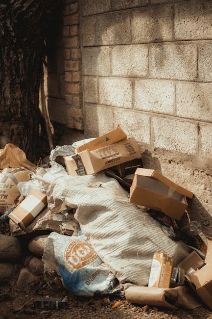 A pile of accumulated waste and packaging materials situated outdoors at the base of a beige brick wall, with a large tree trunk partly visible on the left side. The waste comprises flattened cardboard boxes, some still bearing printed labels, and plastic wrapping, all resting on a collection of small stones and dirt. Among the refuse, there is a white woven sack partially filled and an orange and white branded plastic bag. The scene appears to be an informal rubbish collection point, possibly prepared for removal by a private waste management service such as Rubbish Removal Islington, evident from its positioning against the wall and on the ground, illustrating typical materials handled during on-site clearance or alternative waste disposal methods outside of council collection. The lighting suggests daytime with natural light illuminating the area, emphasizing the textures of the cardboard, plastic, and the rough brick surface behind. The background wall's textured block pattern contrasts with the varied surfaces of the rubbish, emphasizing the disorganized state of the refuse ready for collection or disposal.