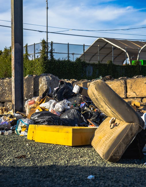 An outdoor scene displaying a collection of discarded rubbish accumulated on a gravel surface, consisting of black plastic garbage bags, a yellow plastic container, a worn car tire, and various small waste items. The black garbage bags appear to be partly torn or loosely tied, revealing some of the waste material inside. The yellow container is lying flat, partially filled with debris. Behind the rubbish, there is a low stone wall constructed from irregularly shaped rough-hewn stones with visible mortar, separating the waste area from a background featuring a dense hedge, tall metal poles, and overhead power lines. Further in the background, a partially visible sports or storage structure with a curved roof is seen, with clear blue sky and a few wispy clouds overhead, indicating daytime weather conditions. This scene may relate to private rubbish collection or on-site clearance activities conducted by companies like Rubbish Removal Islington, highlighting typical elements involved in bulky waste disposal and the importance of proper waste management.