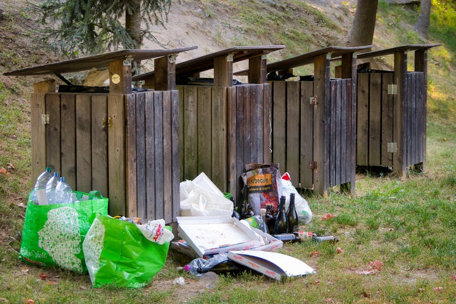 An outdoor scene features four dark-stained wooden waste bins with sloped roofs, arranged in a row on a grassy area with a slight incline. The bins are made of vertical wooden planks, some with visible hinges and locks, and are positioned against a backdrop of trees and shrubbery. In front of the bins, there is a collection of discarded items, including a large green plastic bag filled with plastic bottles, a white pizza box, empty glass bottles, and various pieces of waste such as paper and plastic. The debris is scattered on the grass, some leaning against the bins or lying on the ground. Overhead, natural daylight illuminates the scene, casting soft shadows. The environment appears to be a natural or semi-natural setting, and the presence of waste suggests a situation of improper rubbish disposal, highlighting the importance of proper rubbish removal services such as those offered by Rubbish Removal Islington. This image underscores the need for designated waste management solutions in outdoor spaces to prevent littering and maintain environmental cleanliness.
